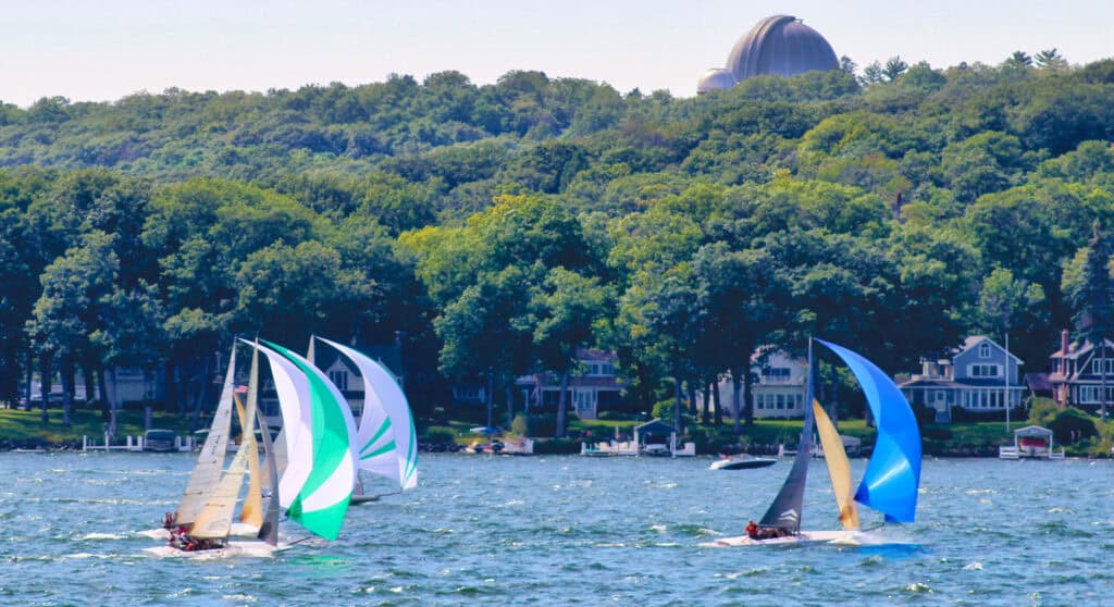 Sailboats racing on Lake Geneva during summer with lush green trees in background.