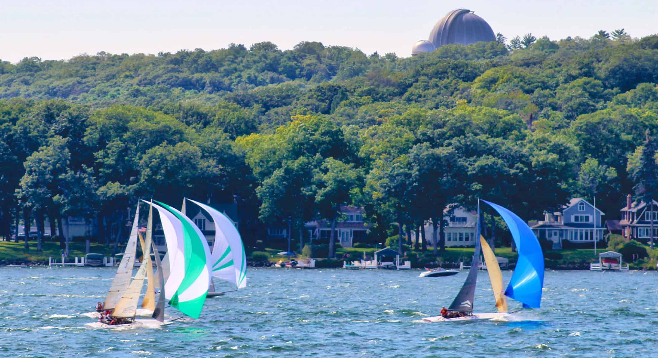 Sailboats racing on Lake Geneva during summer with lush green trees in background.