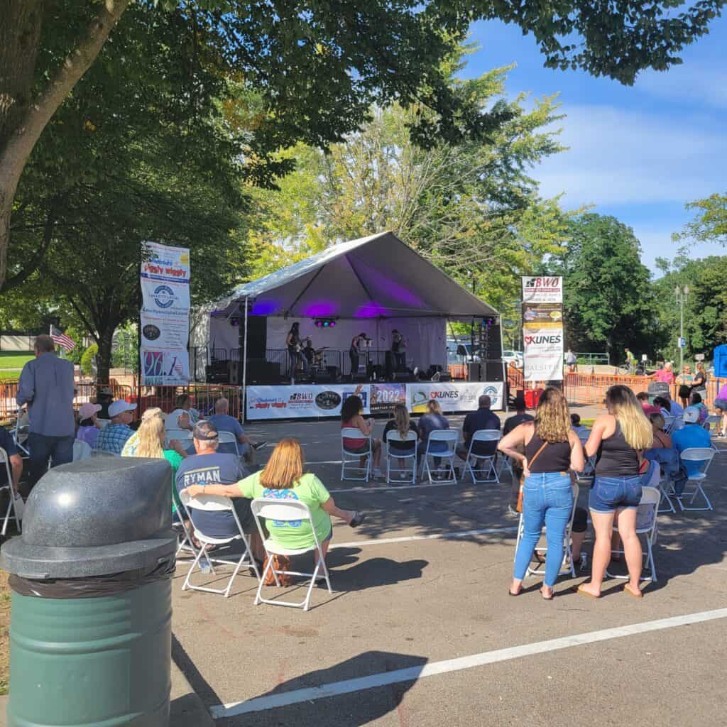 Outdoor live music performance at Lake Geneva with audience and stage under trees.
