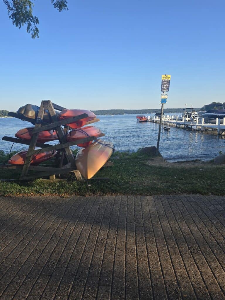 Stacked kayaks on a rack near a calm lake with a dock in the background.
